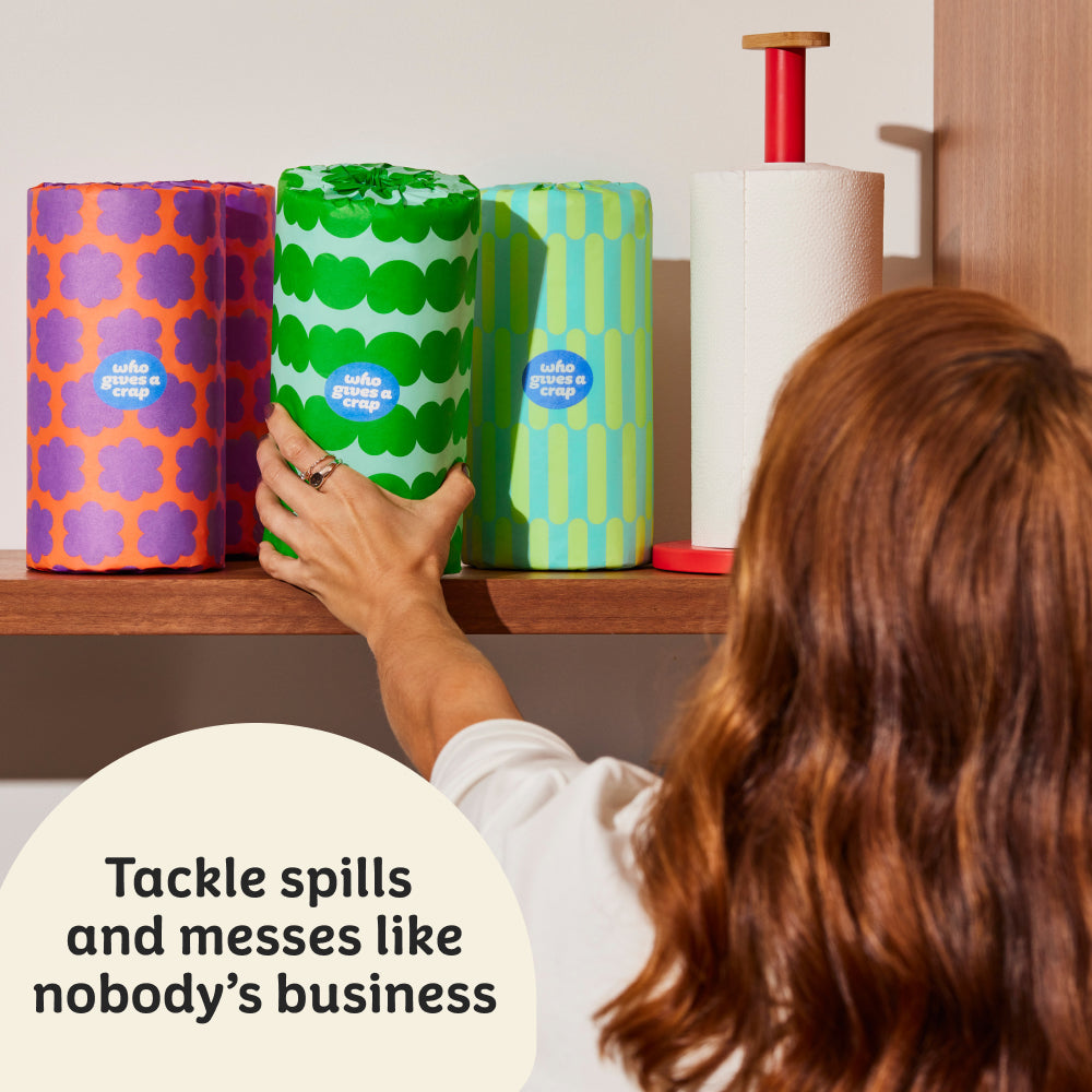 Woman reaches to grab colourful roll of Who Gives a Crap paper towel from kitchen shelf. Sticker on top of image that says ‘tackle spills and messes like nobody’s business’.