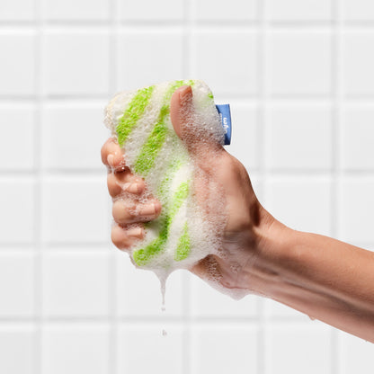 A hand squeezing a lathered Who Gives A Crap green striped scrub sponge against a tiled wall, with soapy water dripping down